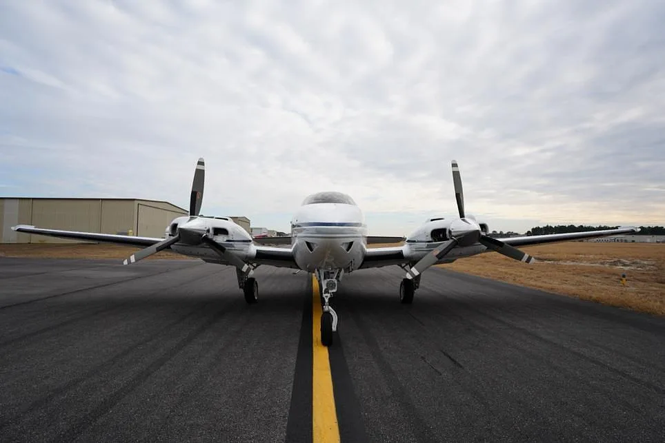 A head-on view of a white twin-engine propeller aircraft with blue and grey stripes on an asphalt runway.