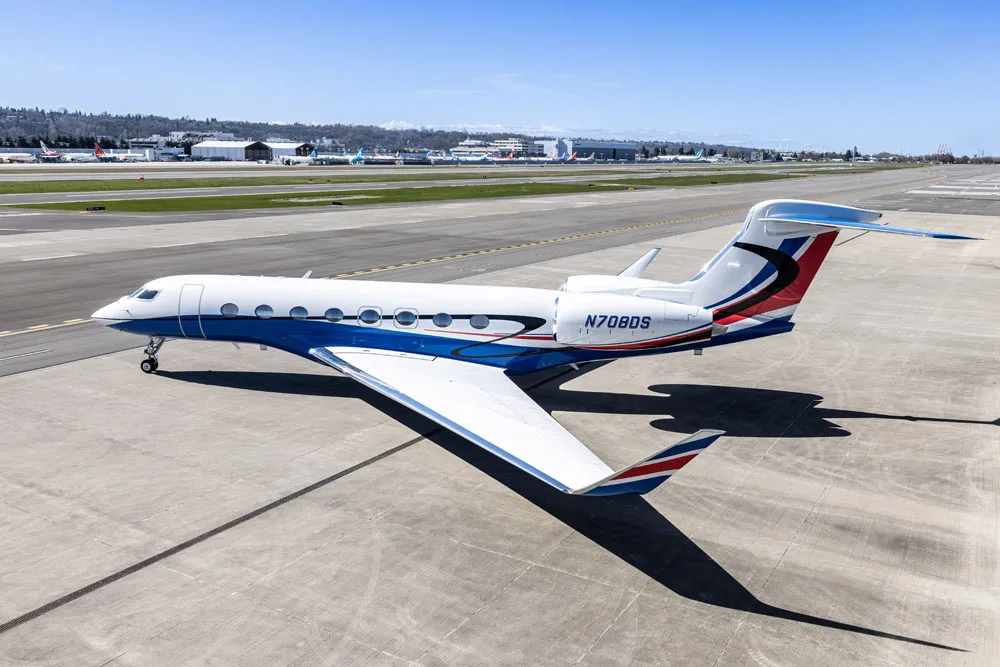 A side profile view of a Gulfstream G650 business jet with a white and blue paint scheme, parked on an airport tarmac.