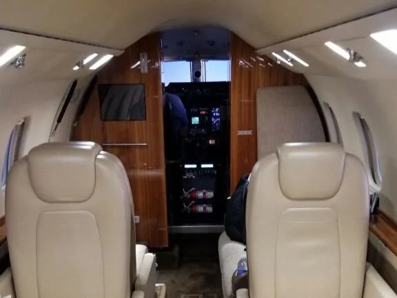 The interior cabin of a private jet with light beige leather seats and wood paneling, looking towards the cockpit.