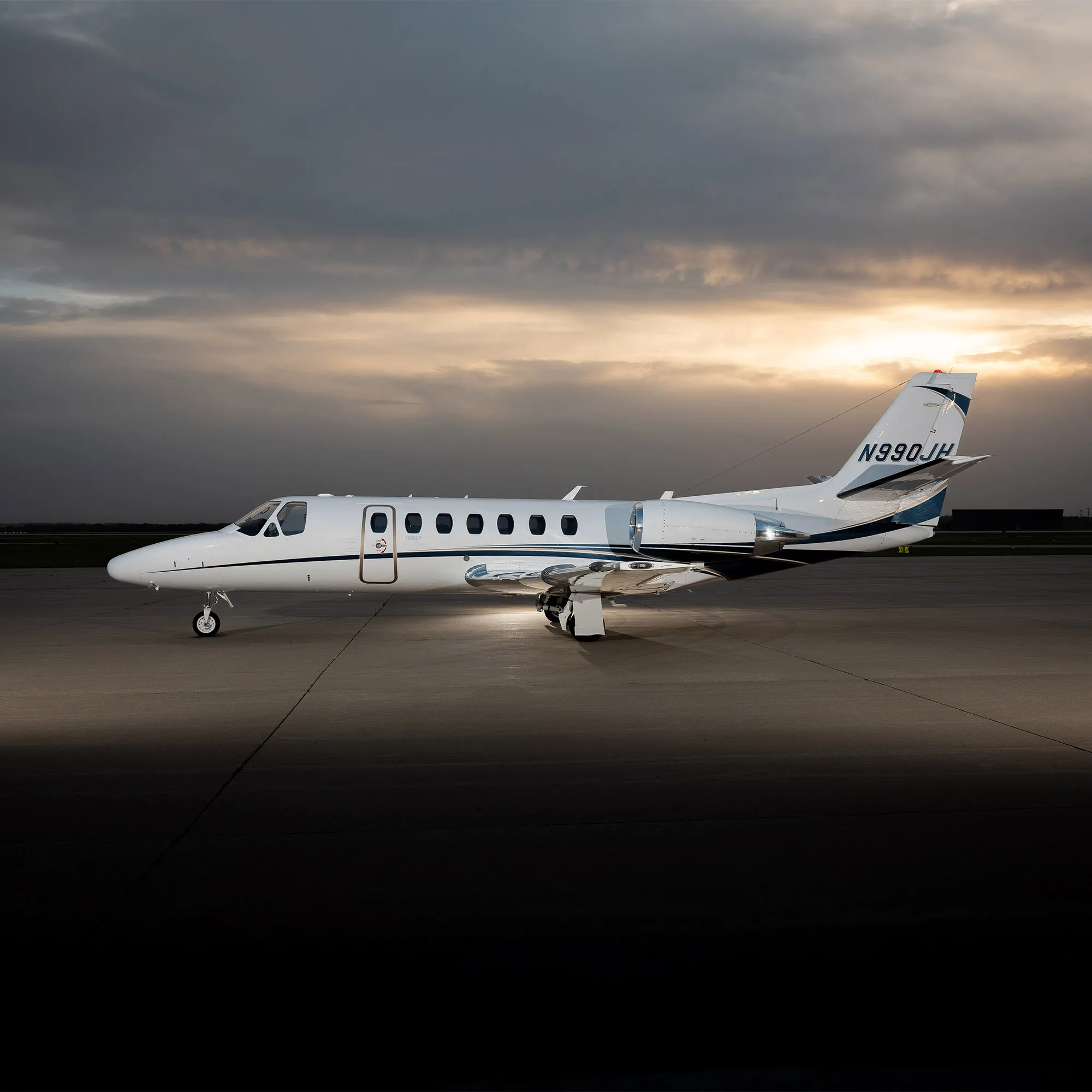 A white Cessna Citation jet with blue and black stripes, registration N990JH, parked on an airfield at sunset.