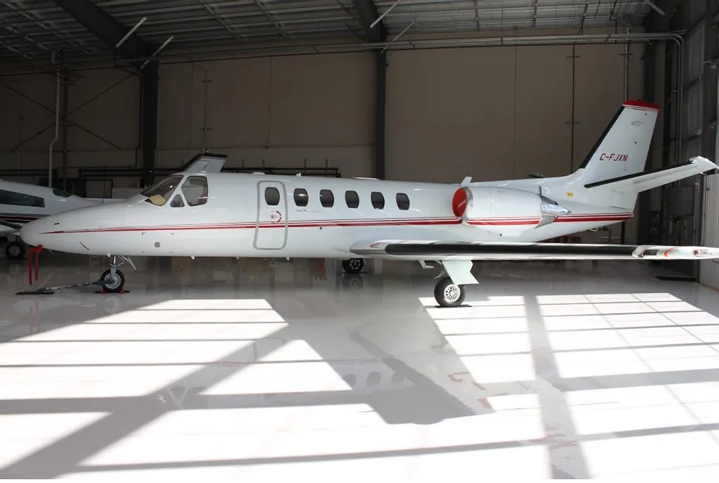 A white Cessna Citation aircraft with red stripes parked in a hangar, viewed from the left side.