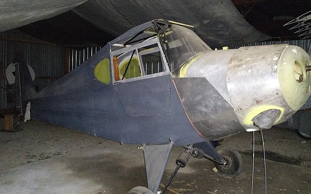 A partially assembled aircraft fuselage with a bare metal nose and grey primer body, seen in a hangar.