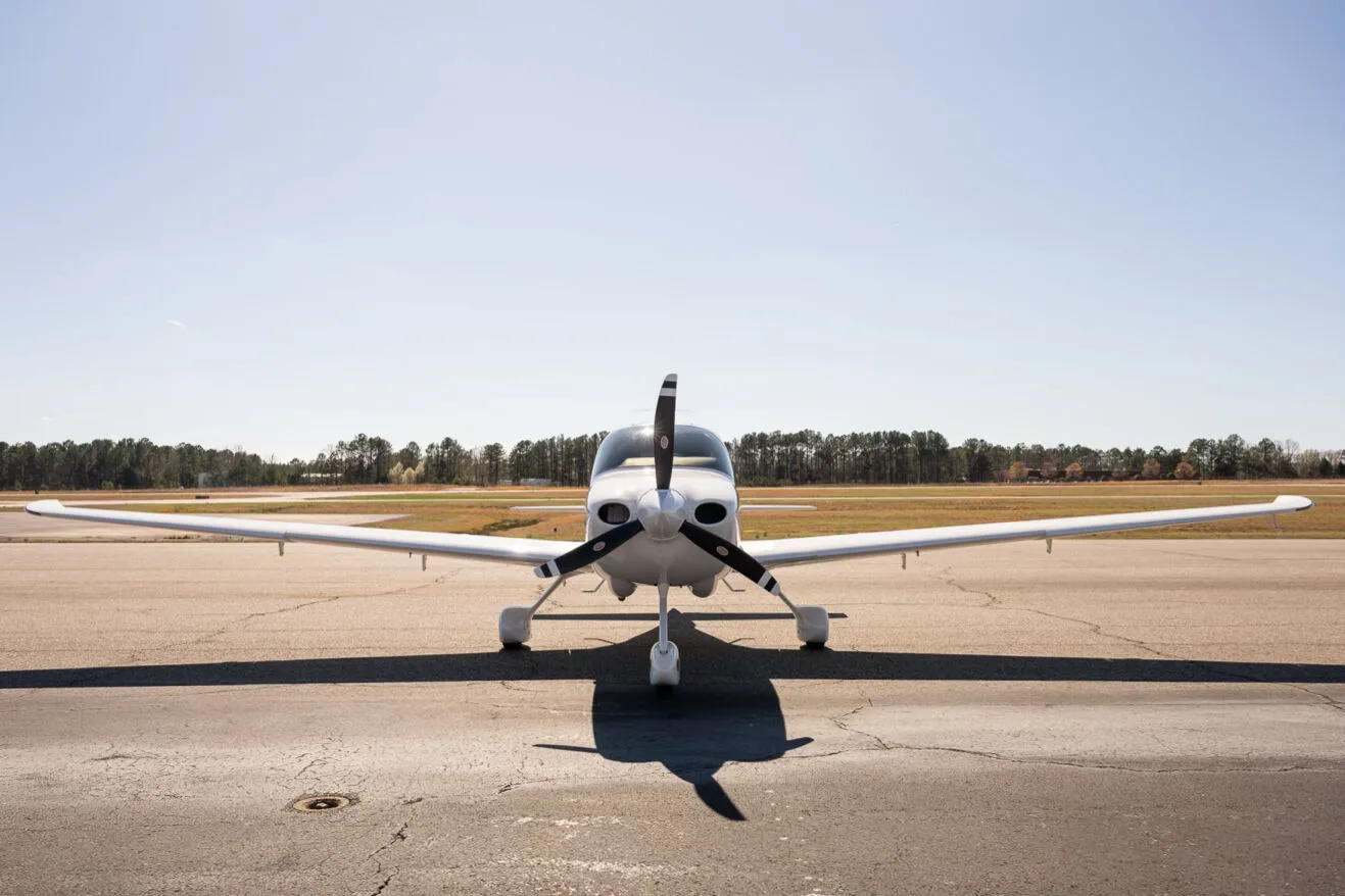 A head-on view of a white Cirrus SR22 aircraft with a three-bladed propeller, parked on an airfield.