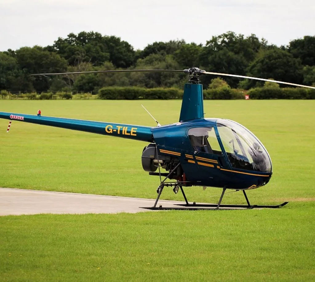 A teal Robinson R22 helicopter with yellow stripes, registration G-TILE, parked on a grassy field.