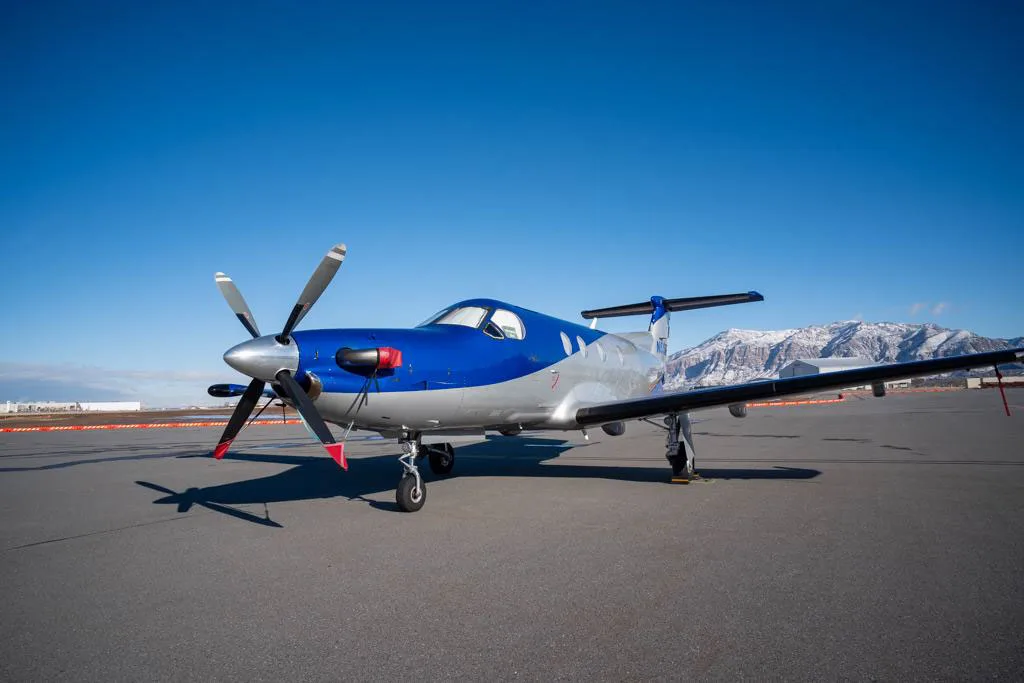 A low-angle front view of a blue and silver Pilatus PC-12 aircraft parked on an airfield under a clear blue sky.