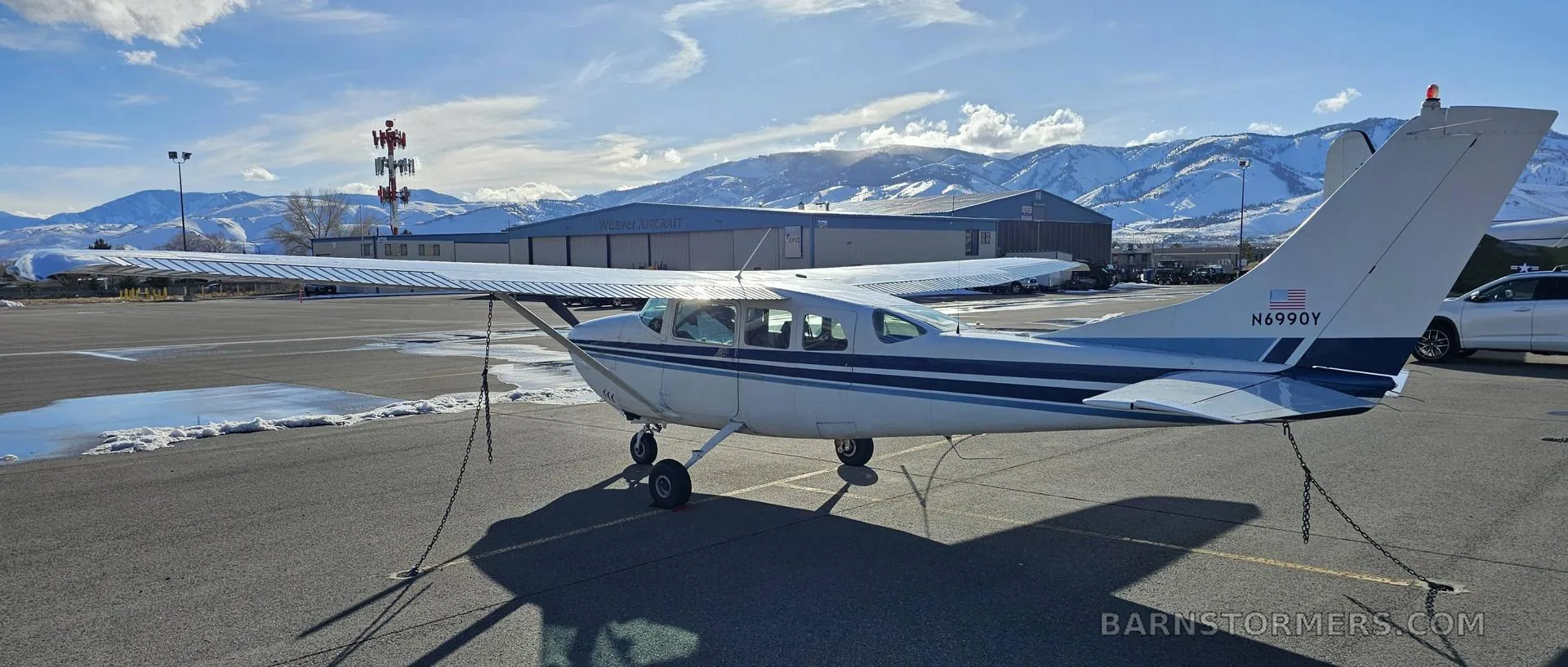A rear-side view of a white and blue Cessna 182 aircraft with registration N6990Y, parked on an airfield with hangars and snowy mountains.