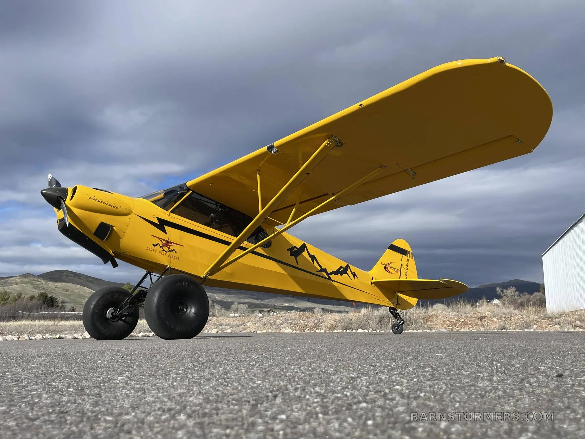 A low-angle side view of a yellow CubCrafters Carbon Cub EX-2 with mountain decals and large tires, parked on an airfield.