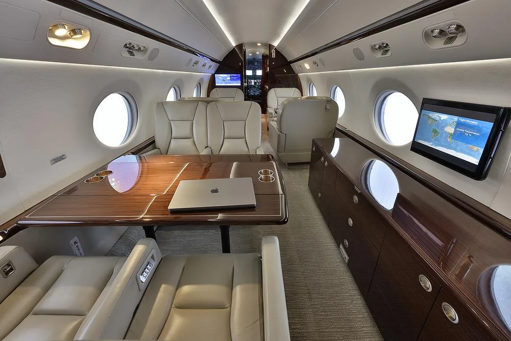 A wide view of a private jet's elegant cabin with beige seating, a large wooden table with a laptop, and wall-mounted entertainment screens.