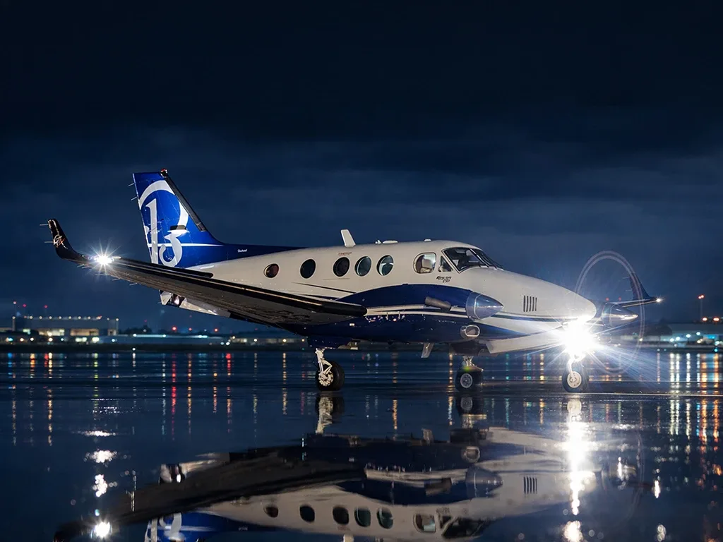 A white and blue twin-turboprop aircraft with a '13' logo on its tail sits on a wet tarmac at night, with landing lights on and city lights reflecting in the water.