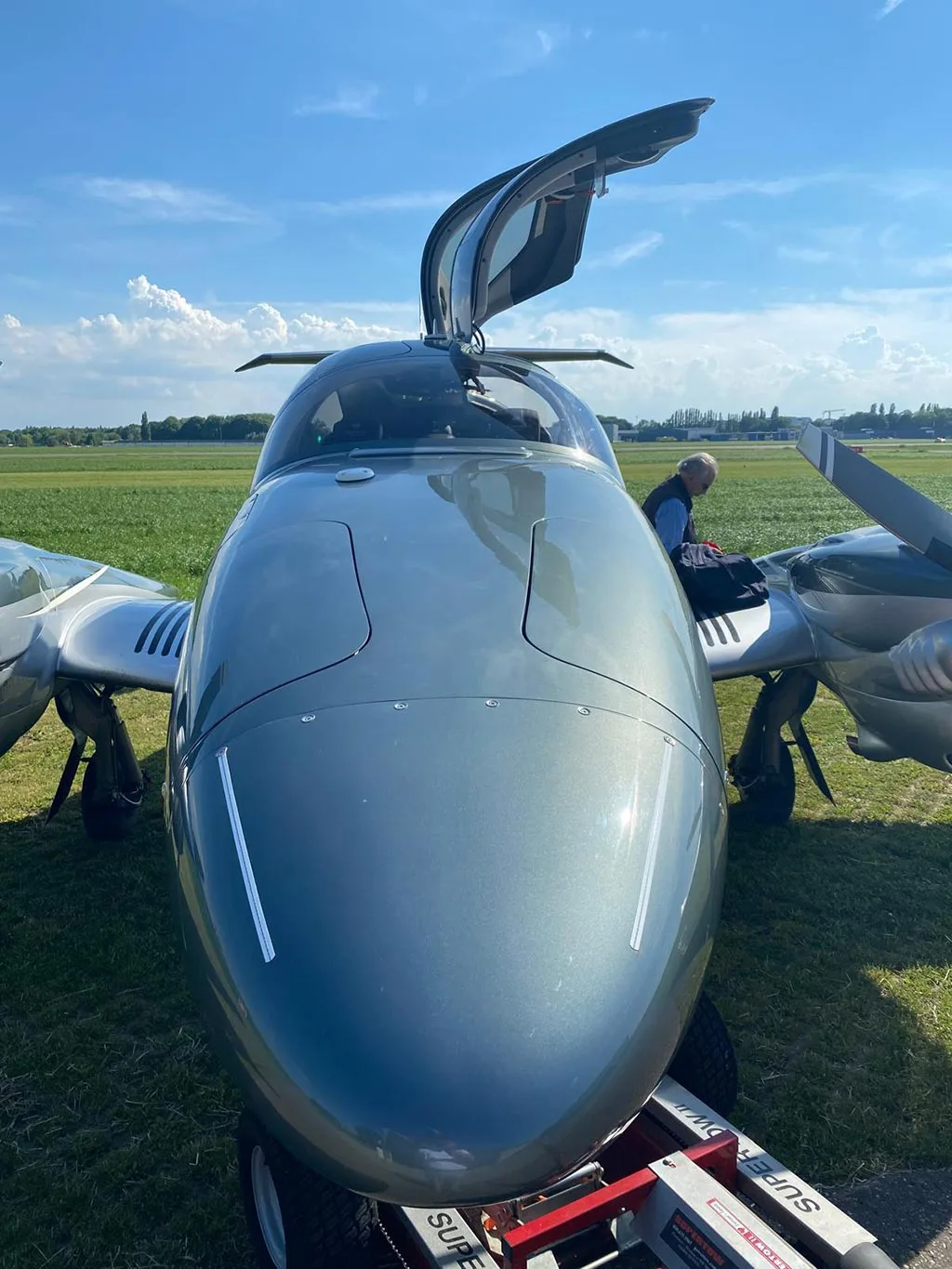 A head-on view of a dark grey Diamond DA62 aircraft with one gull-wing door open, being moved by a tug on a grassy field.