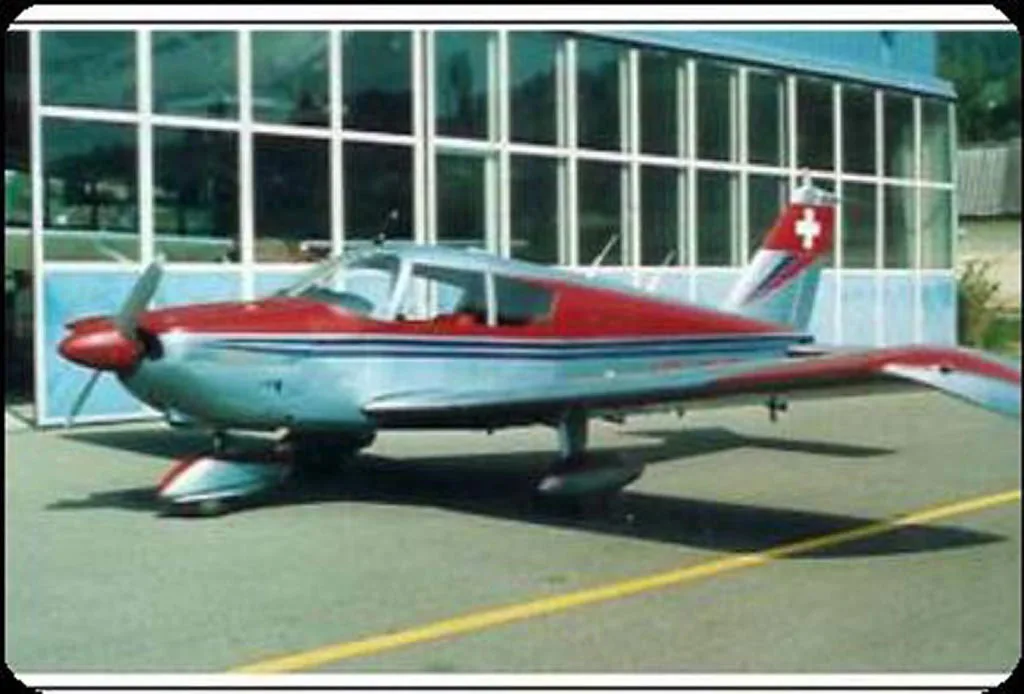 A red and silver single-engine propeller aircraft with a Swiss flag on its tail, parked on an tarmac in front of a building.