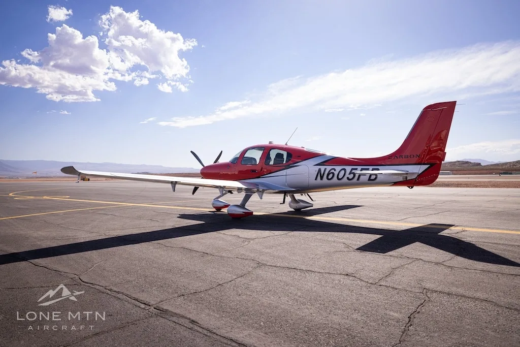 A rear-quarter view of a red and silver Cirrus SR22T aircraft (N605FB) with its tail facing the viewer, on an airfield.