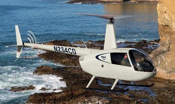 A white Robinson R44 Raven helicopter with registration N234CD hovers low over a rocky coastline with ocean waves.