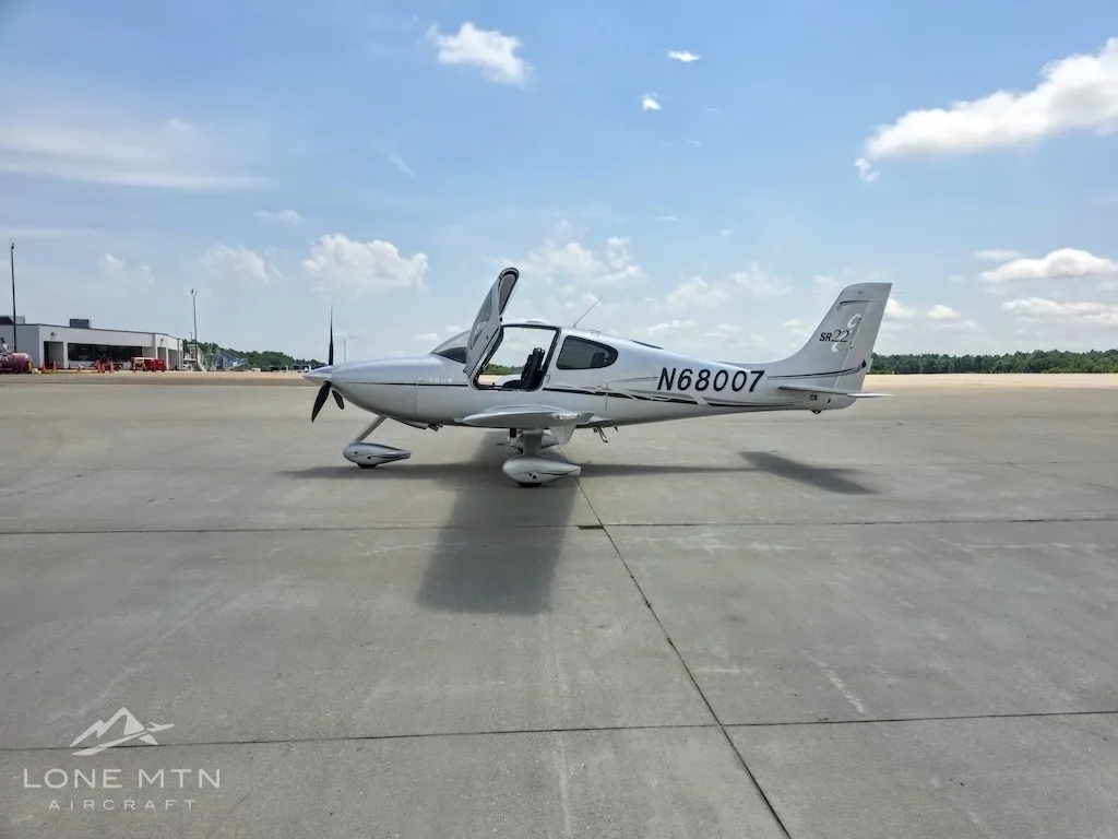 A silver Cirrus SR22 aircraft (N68007) with its gull-wing doors open, parked on an airfield, viewed from the front-right.