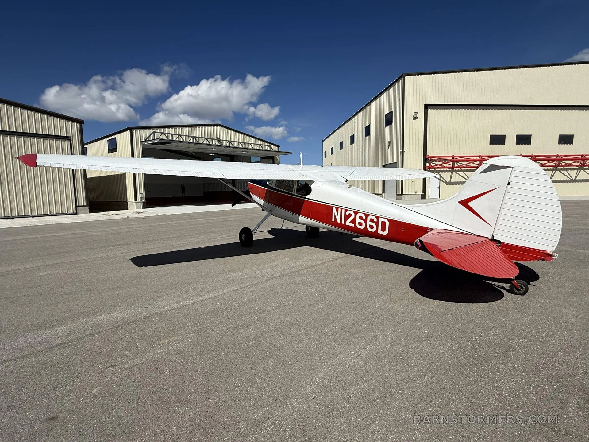 A red and white Cessna 170B (N1266D) is parked on an airfield, viewed from the rear-right, with hangars and a blue sky.