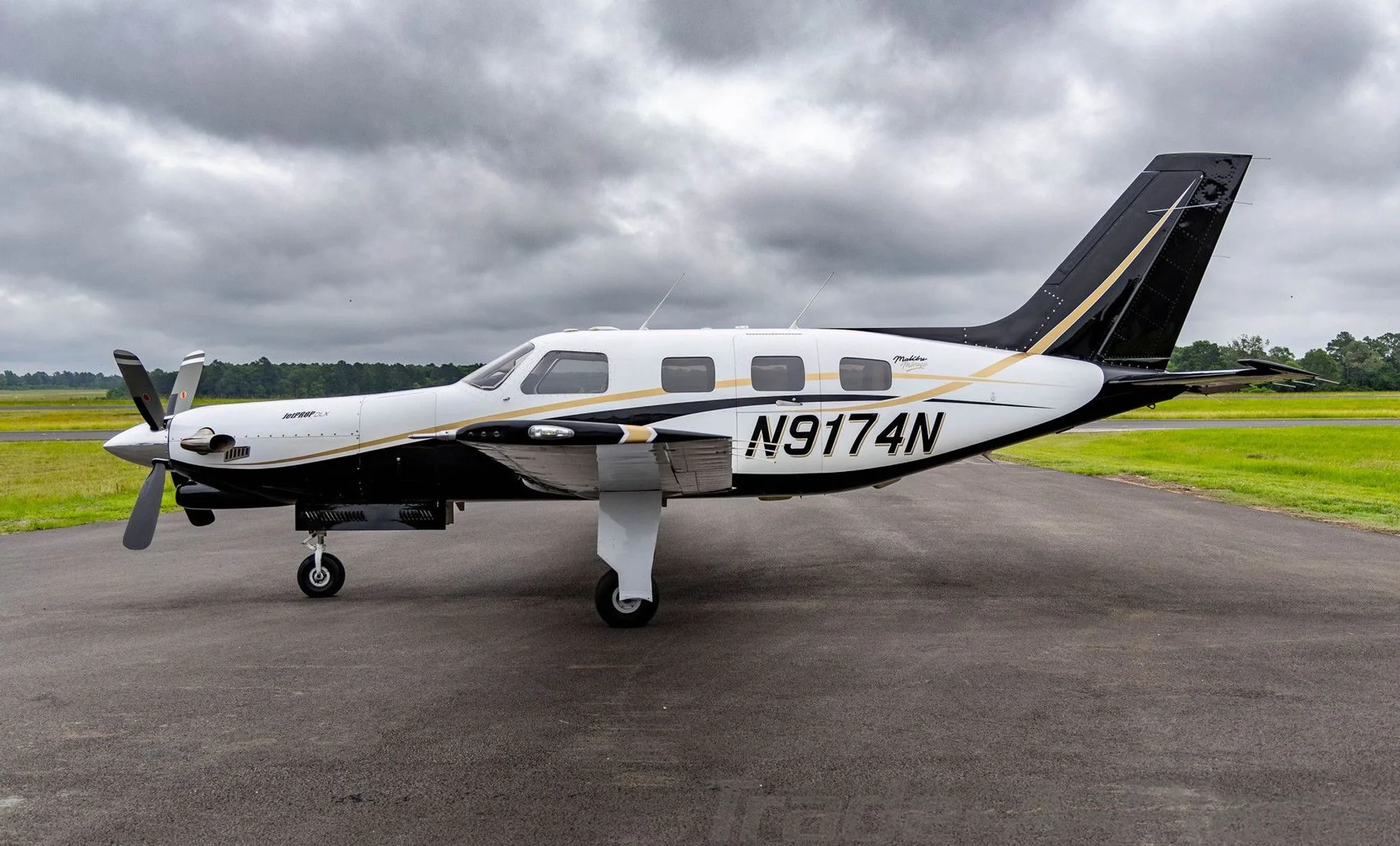A white and black Piper Malibu Mirage (JetPROP DLX) N9174N with gold accents parked on a tarmac under a cloudy sky.