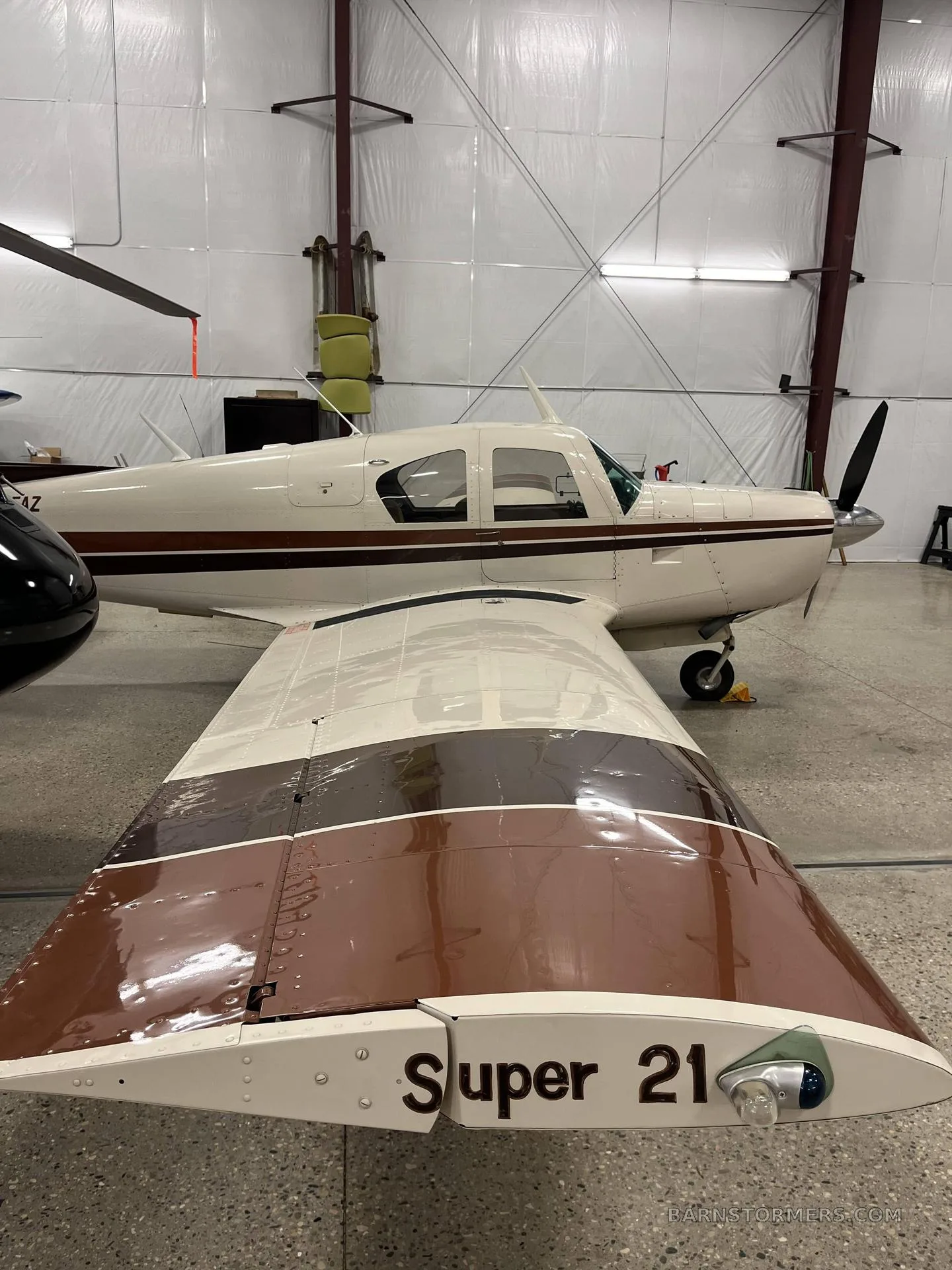A cream and brown single-engine propeller aircraft (Beechcraft Debonair "Super 21") is shown from the rear-right in a hangar, highlighting the wing.