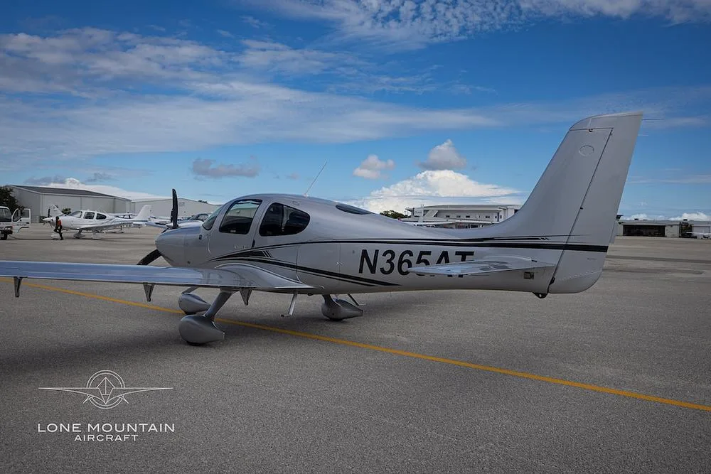 A silver Cirrus SR22 aircraft with black and white stripes, viewed from the rear-left on an airfield.