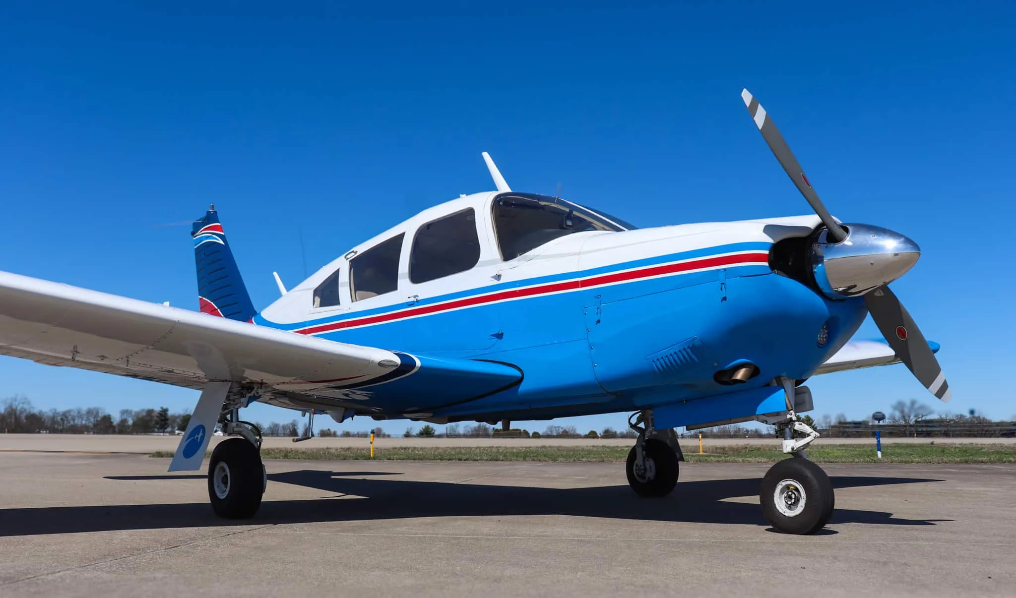 A low-angle front-side view of a blue and white Piper Cherokee aircraft with red stripes on an airfield.