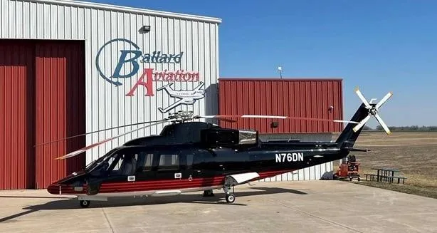 A black and red helicopter, N76DN, is parked on a tarmac in front of a white and red hangar.