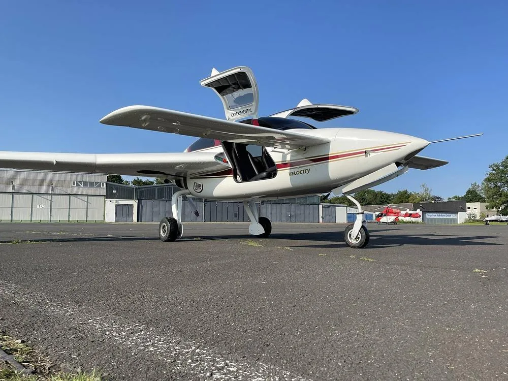A white Velocity Elite aircraft with red and gold stripes, registration N96KP, parked on a tarmac with its gull-wing doors open.