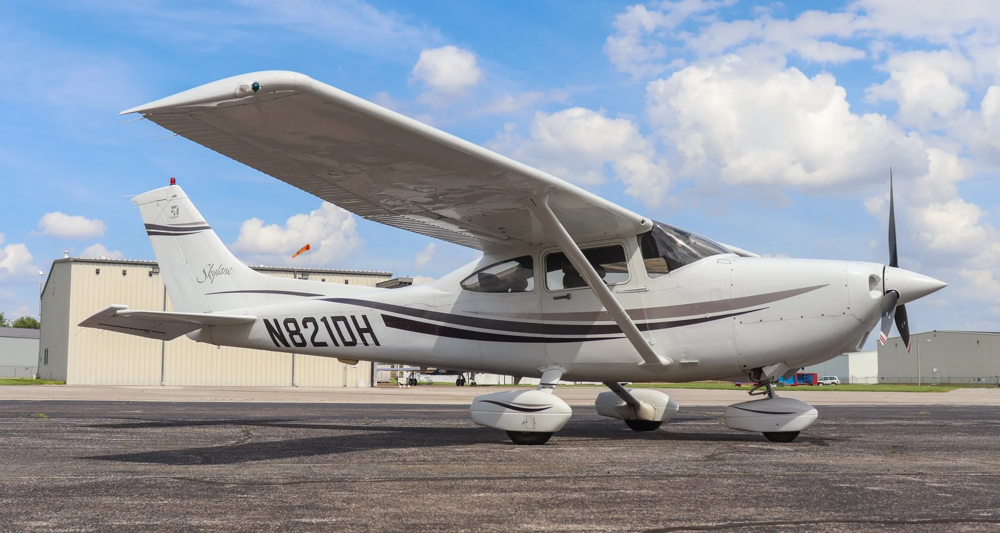 A white Cessna Skylane N821DH with dark grey and brown stripes is parked on an asphalt tarmac under a partly cloudy sky.