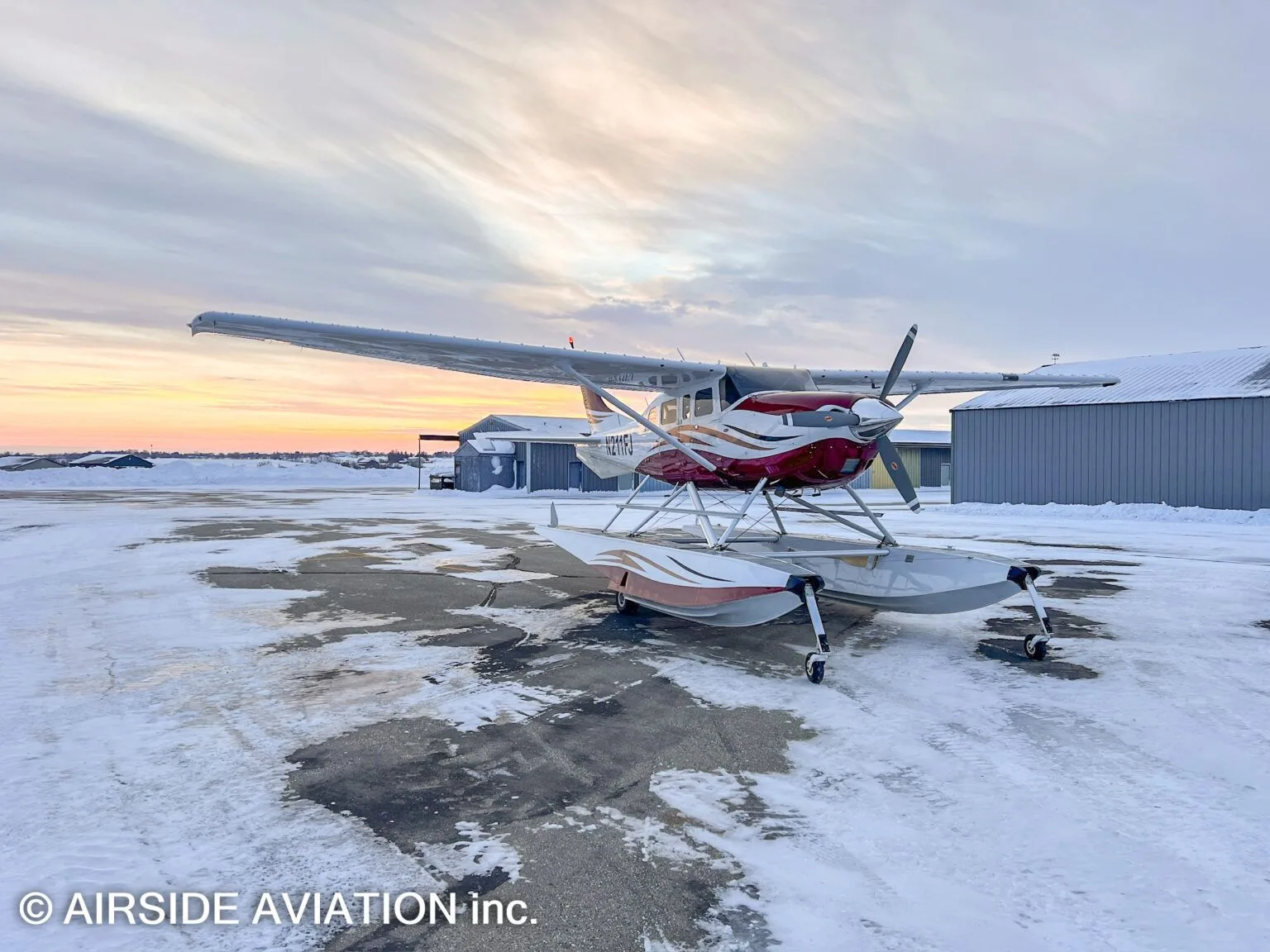 A red and white floatplane, N211FJ, is parked on a snowy tarmac at sunset with hangars in the background.