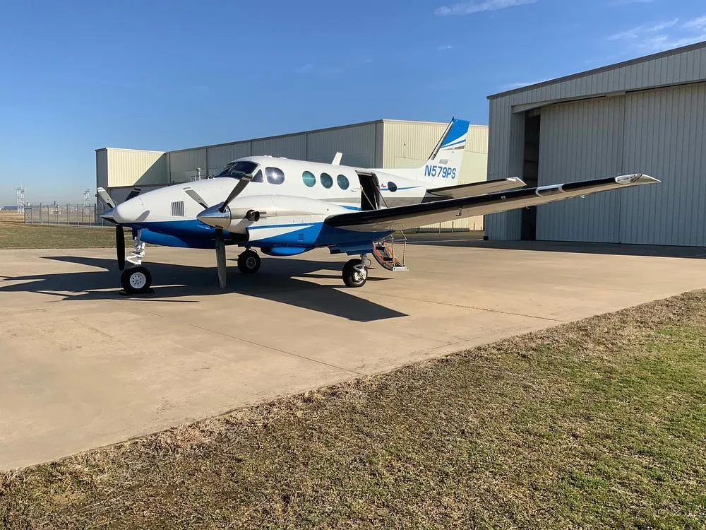 A white and blue twin-engine turboprop aircraft, N579PS, parked on an airfield next to hangars.