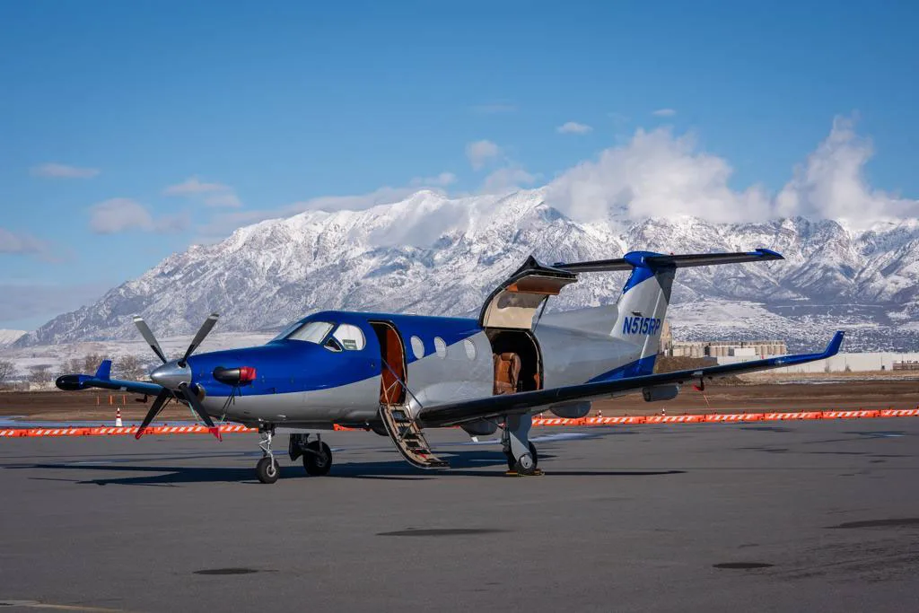 A blue and silver Pilatus PC-12 aircraft with its main door open, parked on an airfield with snowy mountains in the background.