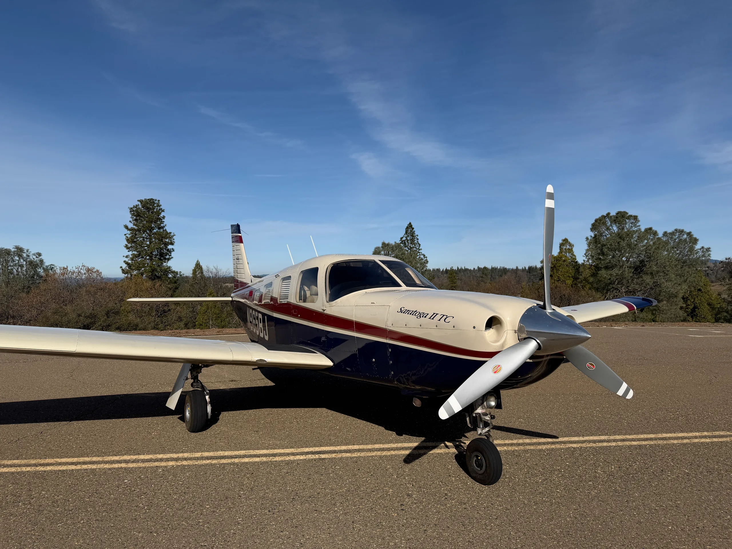 A Piper Saratoga II TC aircraft with a cream, blue, and red paint scheme parked on an asphalt surface with trees in the background.