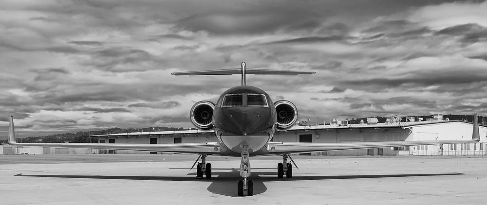 A black and white head-on view of a business jet, likely a Gulfstream G650, parked on an airfield with buildings in the background.