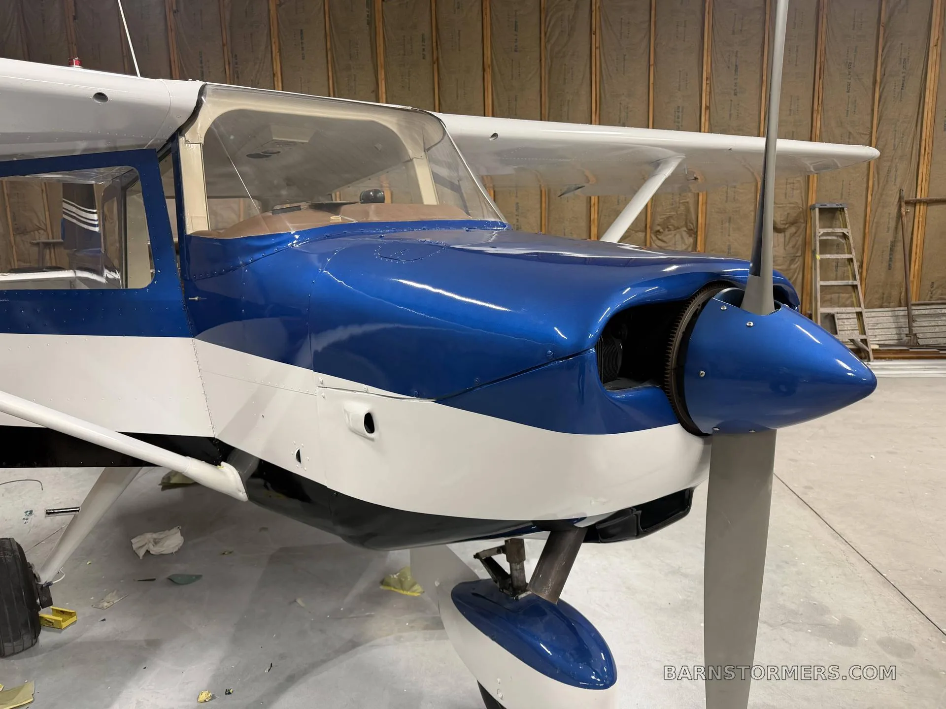 A blue and white single-engine aircraft, likely a Cessna, is shown from the front-right in a hangar.