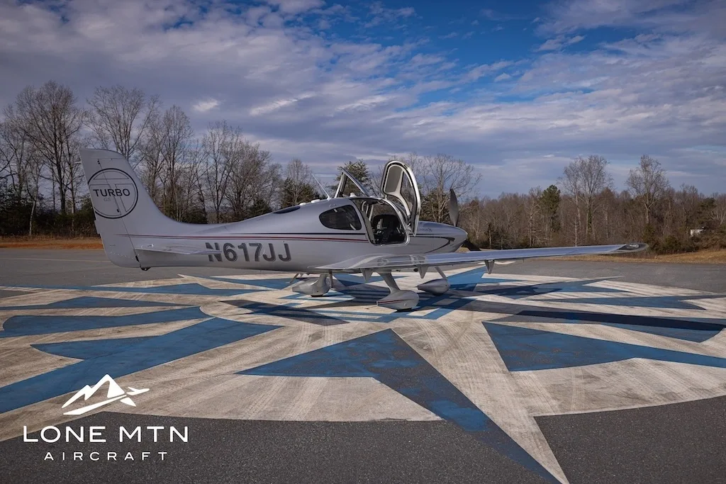 A silver Cirrus SR22T aircraft, N617JJ, with its doors open, parked on an airfield with trees in the background.