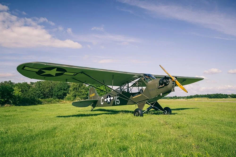 A Piper L-4 Grasshopper in olive drab military paint scheme with US Army Air Forces markings, parked on a grassy airfield under a partly cloudy sky.