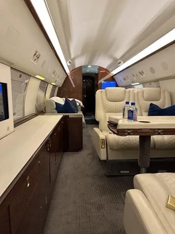 A view inside the private jet cabin shows cream-colored quilted leather seats, a table, and dark wood paneling.