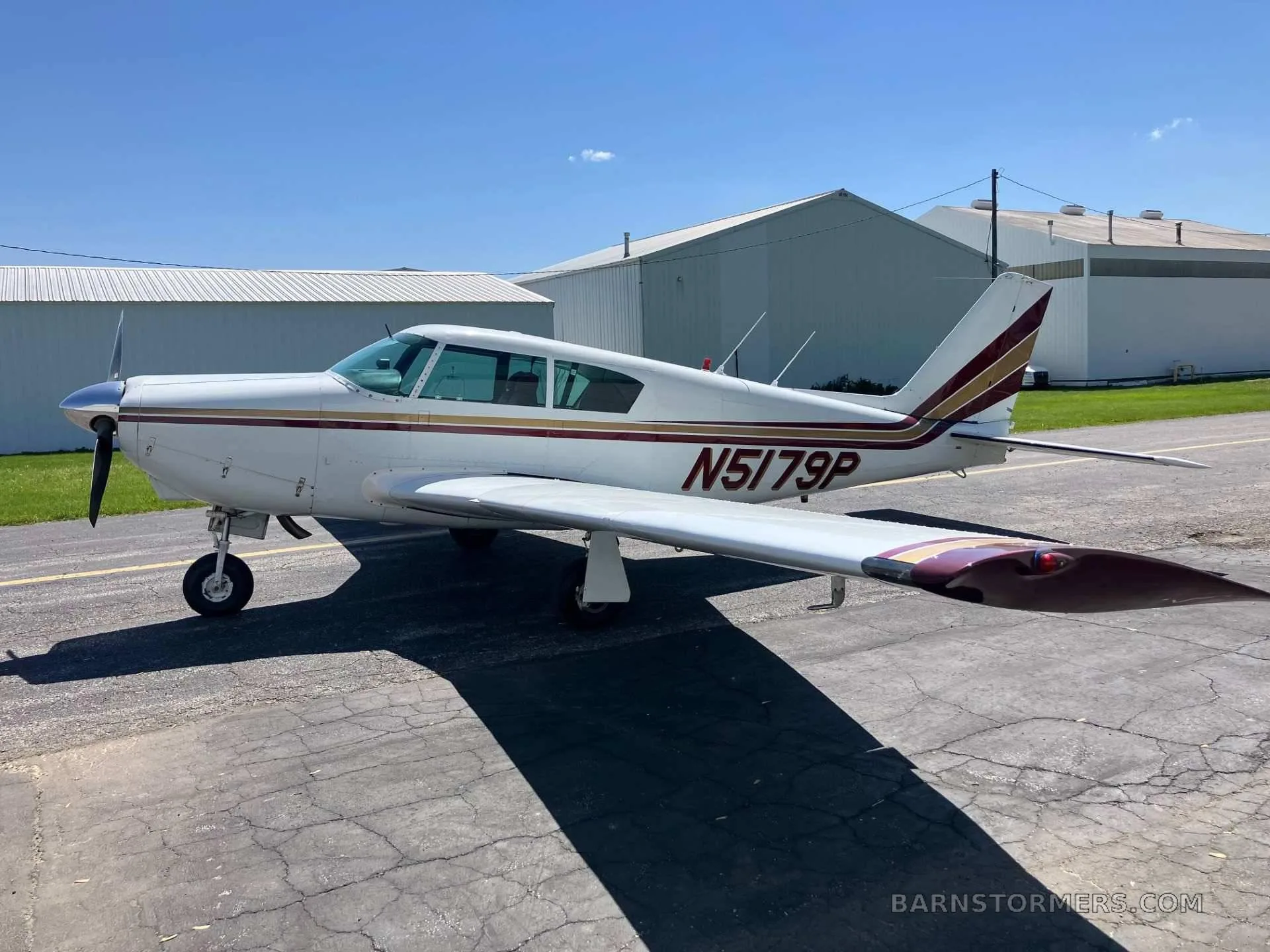 A white single-engine aircraft with red and gold stripes, registration N5179P, parked on an asphalt ramp next to hangars under a clear blue sky.