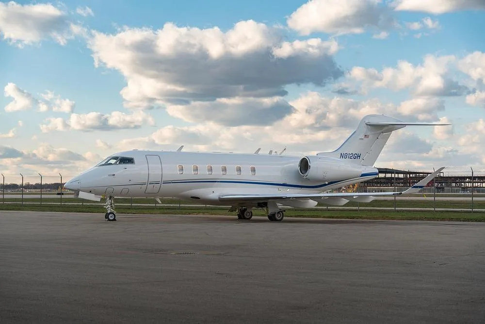 A white Bombardier Challenger 350 business jet with blue stripes on the tarmac under a cloudy sky, seen from the side.