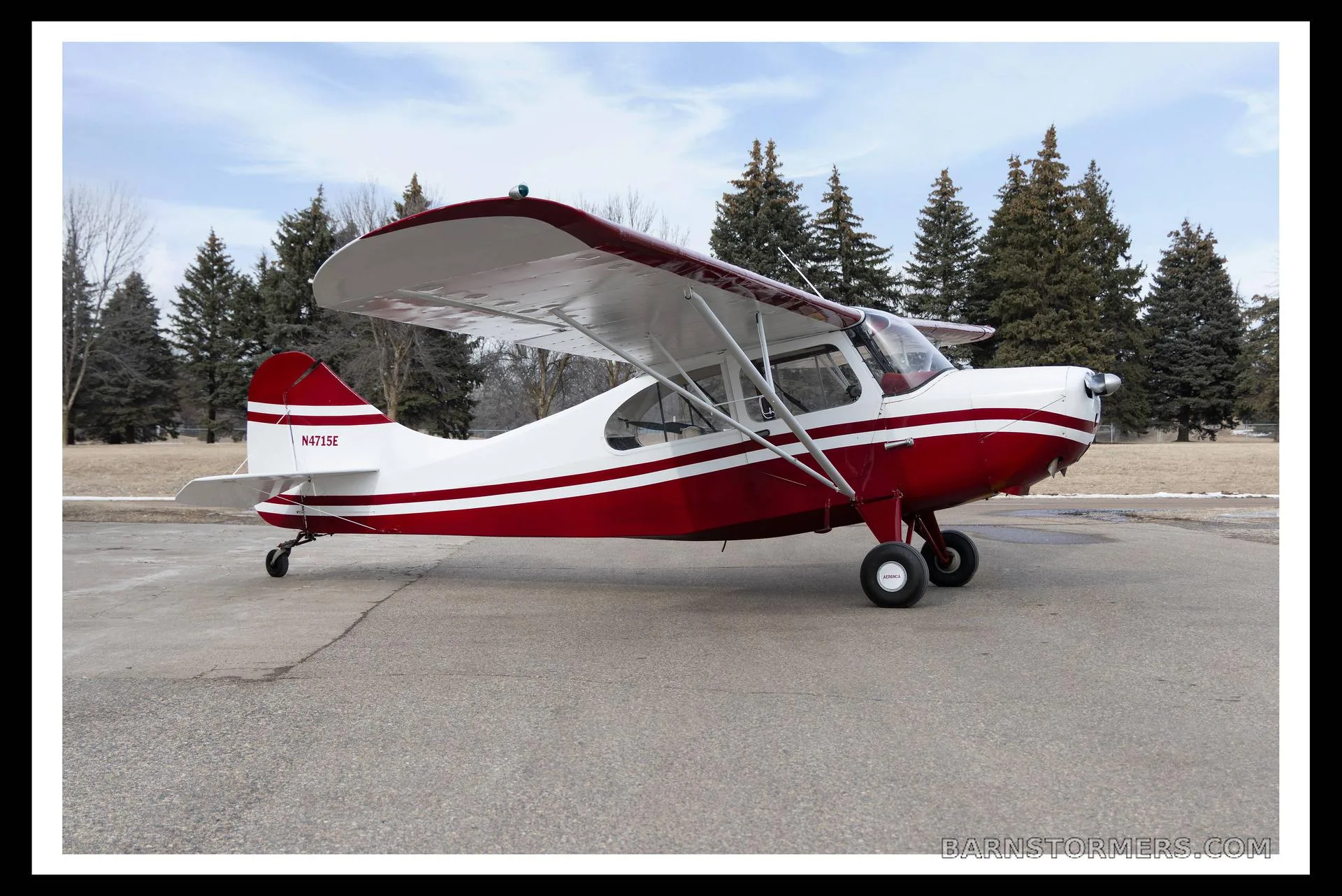 A red and white single-engine taildragger aircraft with registration N4715E, parked on an asphalt surface with pine trees in the background.
