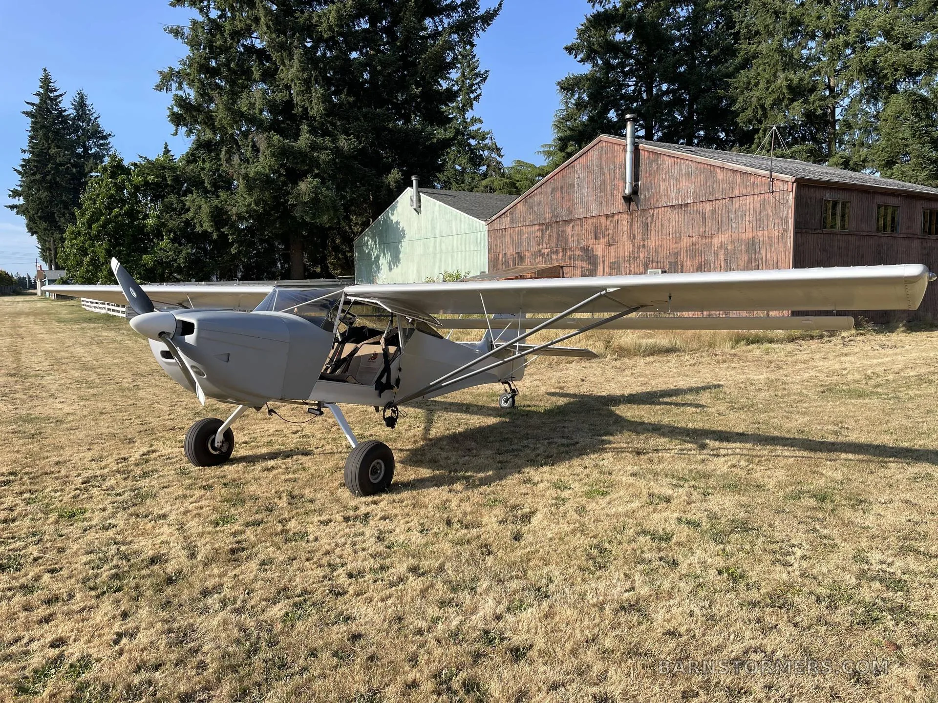 A light grey single-engine aircraft with a high wing and large tires is parked on dry grass with two rustic buildings and trees in the background.