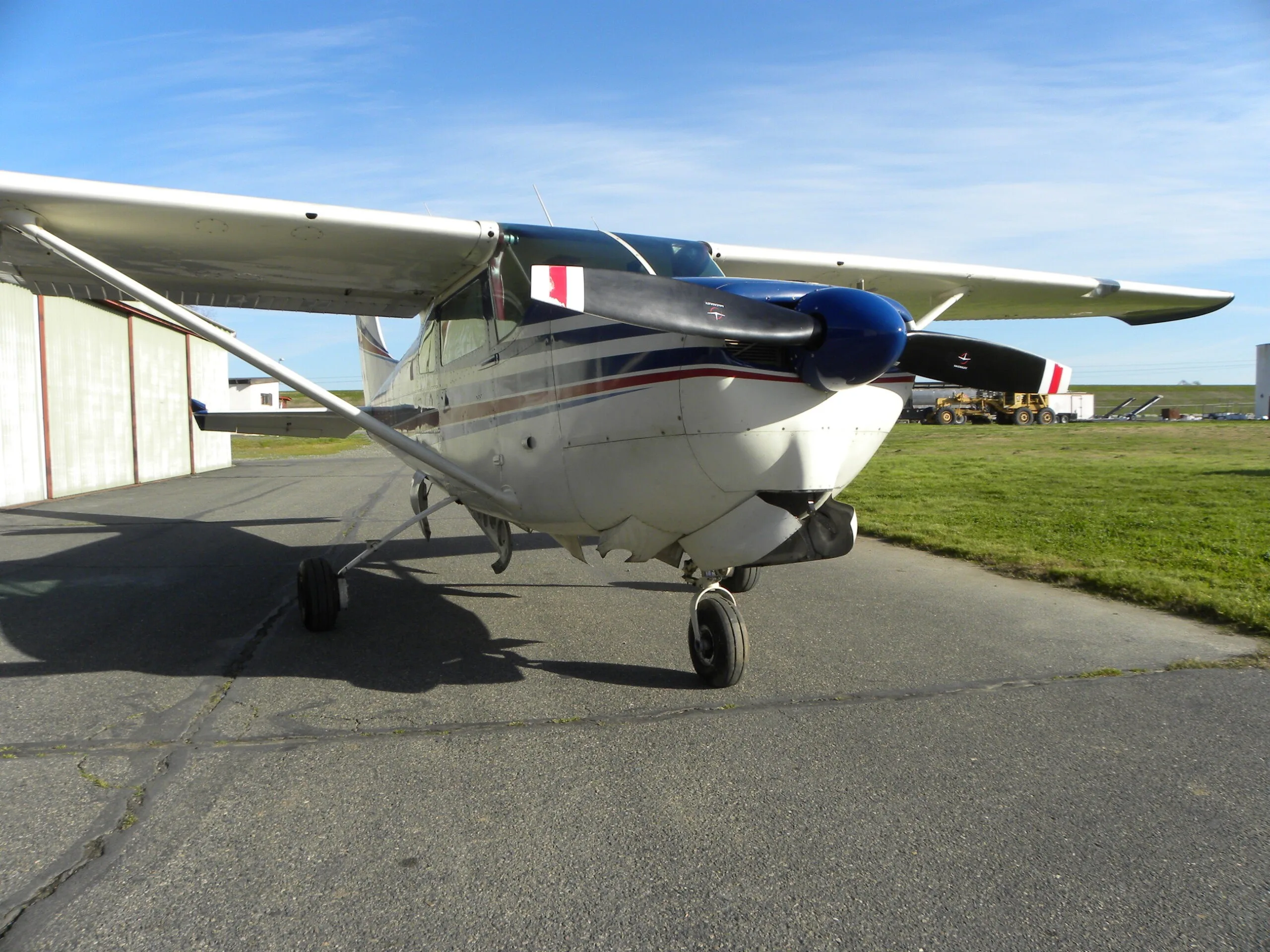 A front view of a white Cessna 182 with blue and red stripes on a tarmac next to a hangar.