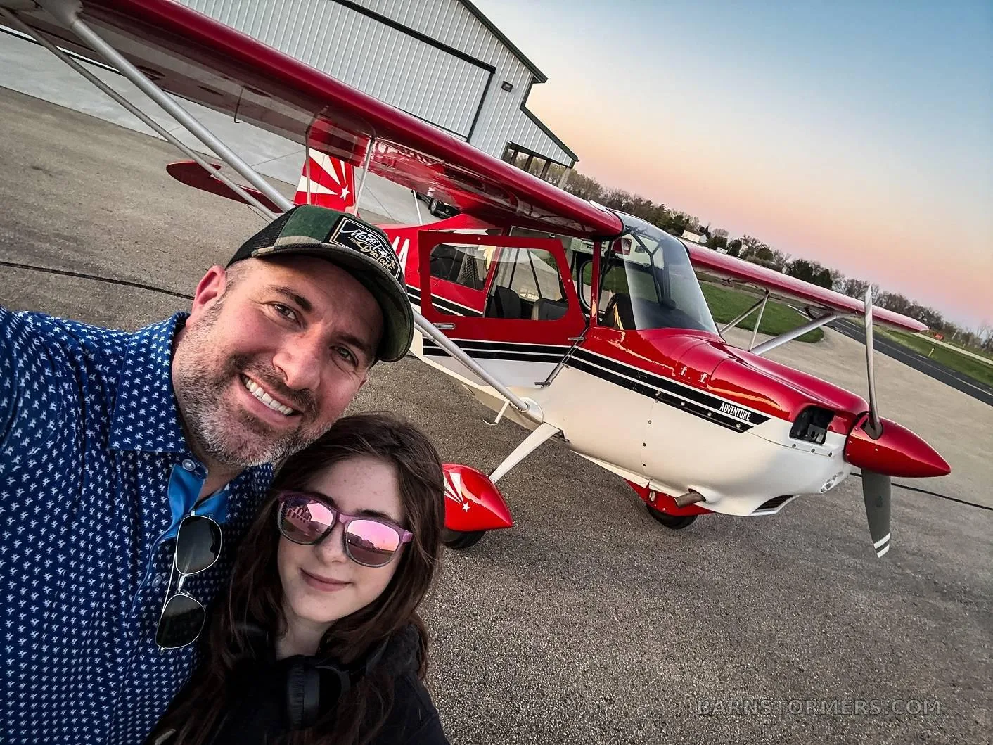 A man and a girl smile for a selfie in front of a red and white single-engine aircraft on an airfield at sunset.