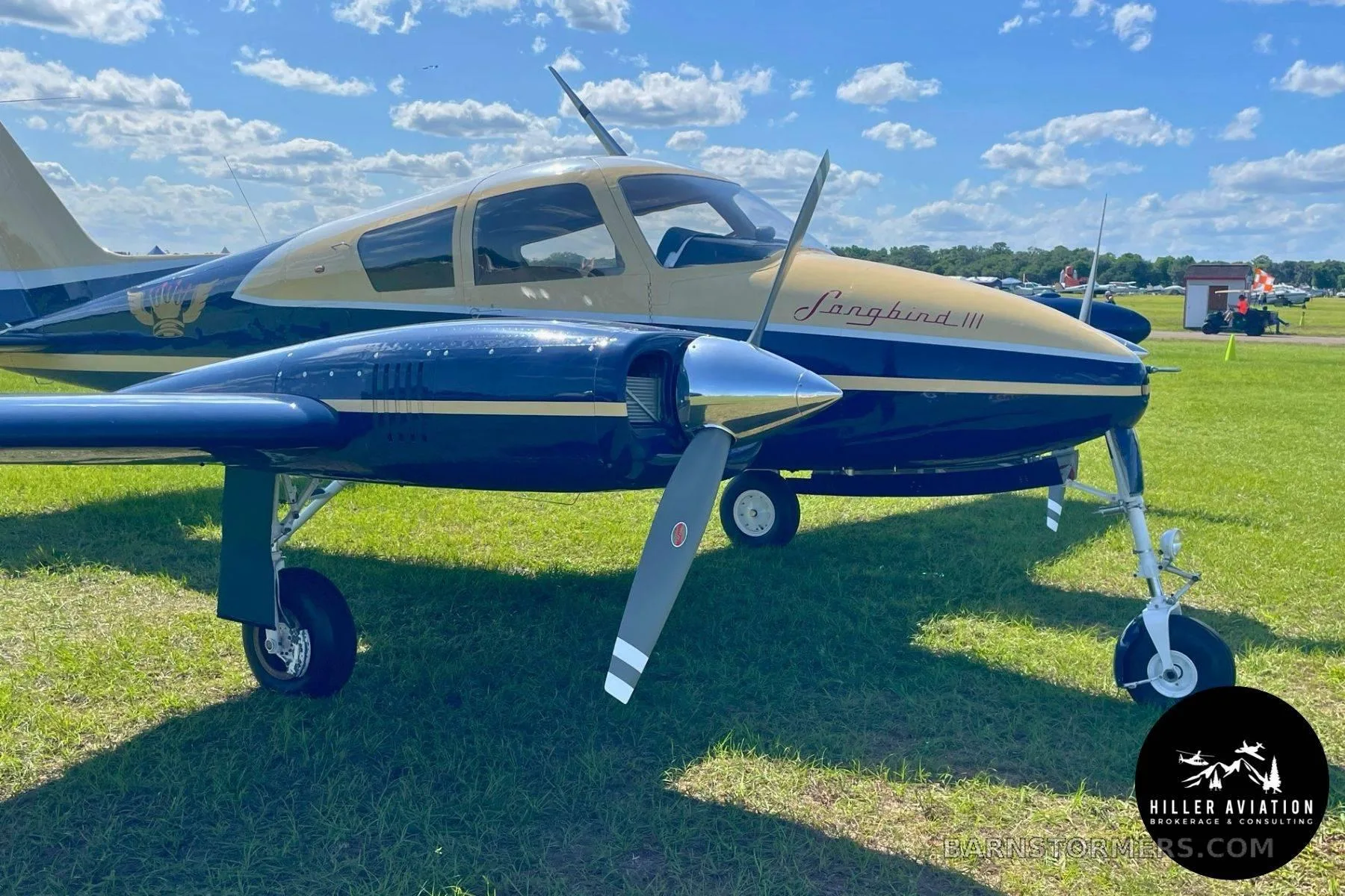 A close-up view of the front of a blue and gold twin-engine aircraft, "Longbird III", parked on a grassy field.