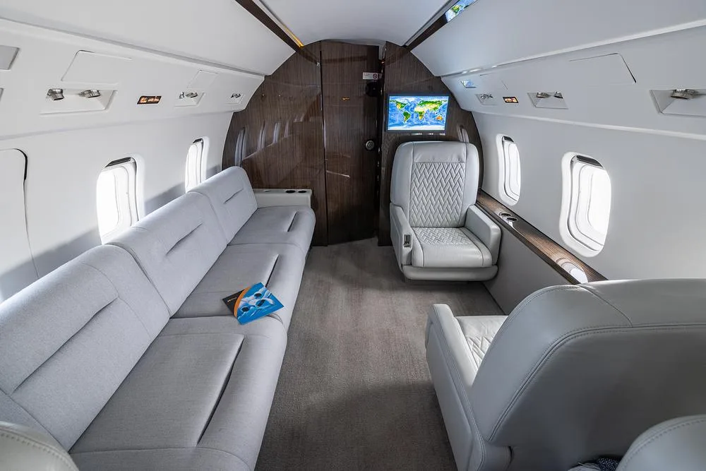 The rear section of a business jet's cabin features a long light gray sofa and an individual seat, with a world map displayed on a screen.