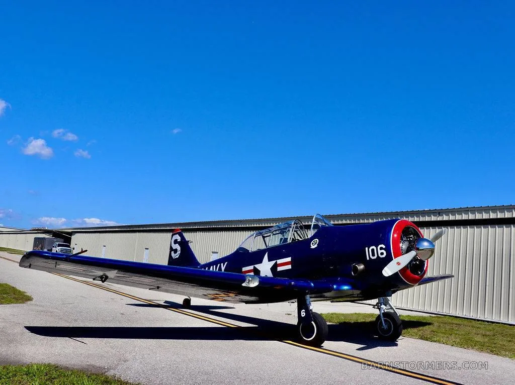 A dark blue vintage military trainer aircraft with "NAVY" and "106" markings is parked on an airfield with hangars in the background.