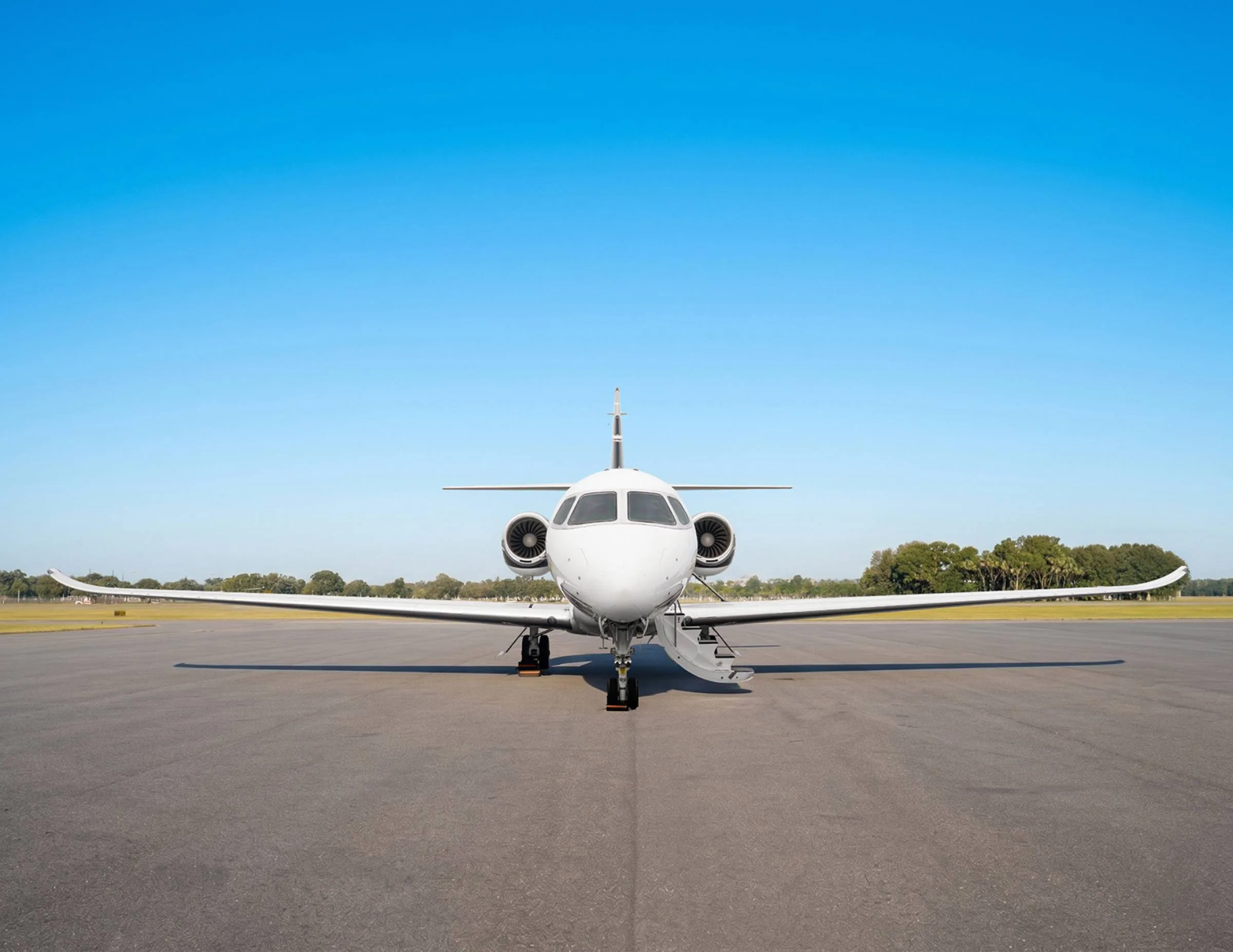 A white business jet with its wings extended is parked on an asphalt runway, viewed directly from the front under a clear blue sky.