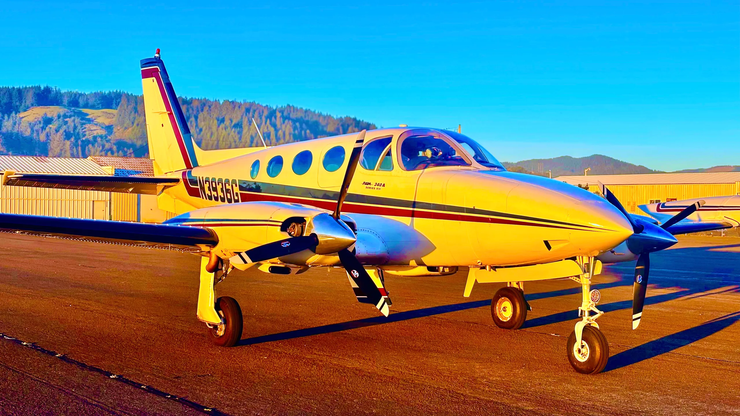 A white and cream twin-engine Cessna 340 aircraft with red and blue stripes parked on an airfield at sunset.