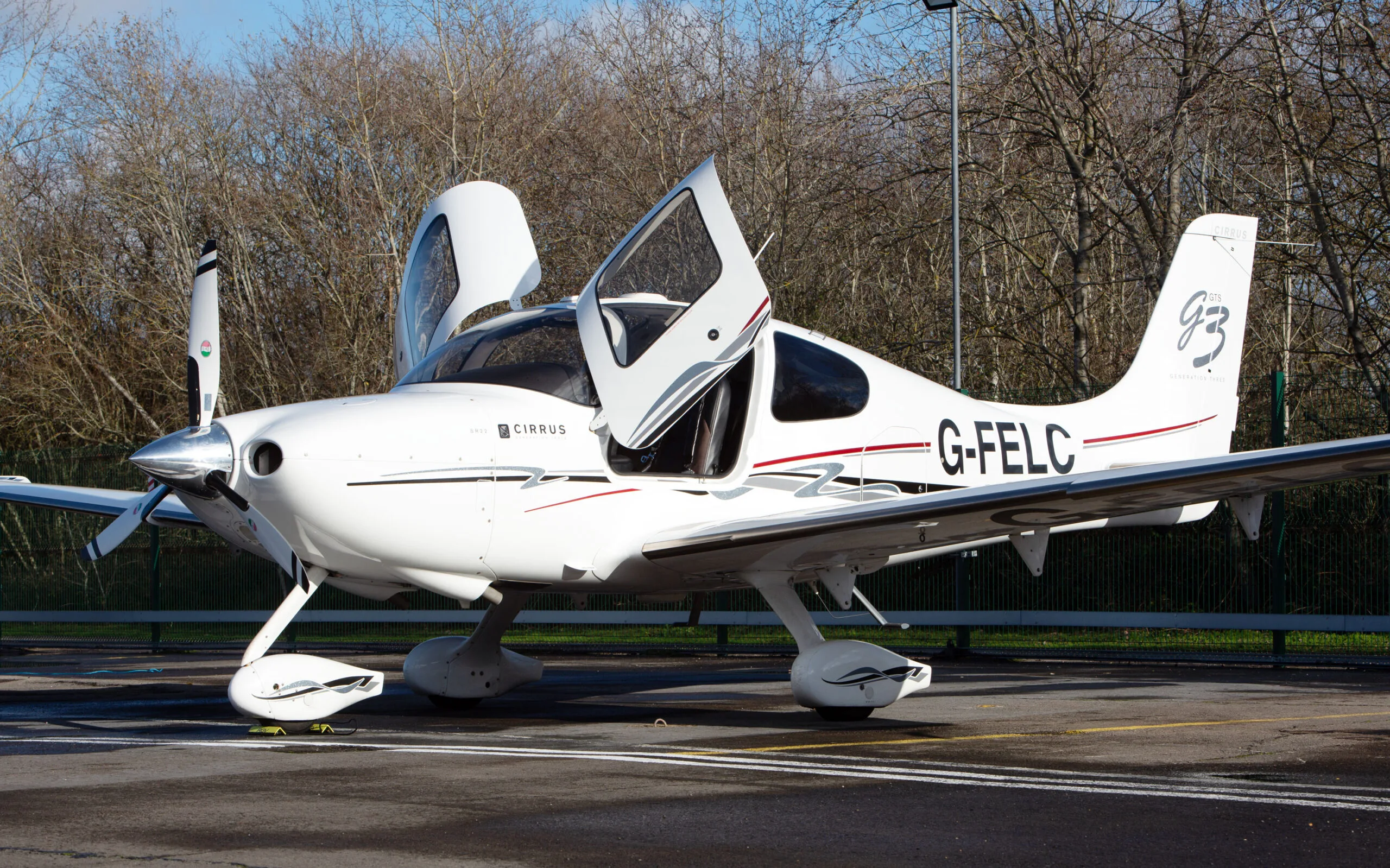A white Cirrus SR22 G3 aircraft with red and black stripes, registration G-FELC, parked on a tarmac with its gull-wing doors open, viewed from the front-right.