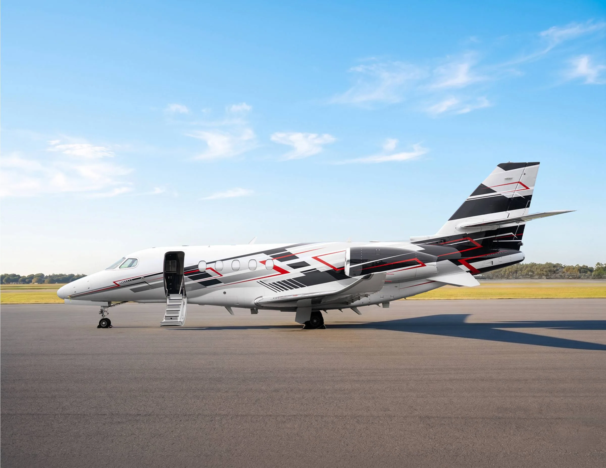 A white business jet with black, red, and grey geometric livery is parked on an asphalt runway with its door open and stairs extended, under a blue sky.