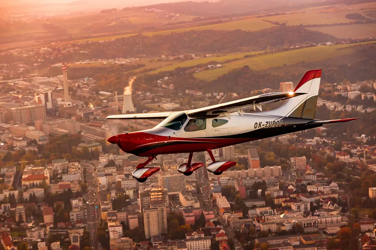 A red and silver Bristell B23 light aircraft with registration OK-ZUR77 flies over a city at sunset.