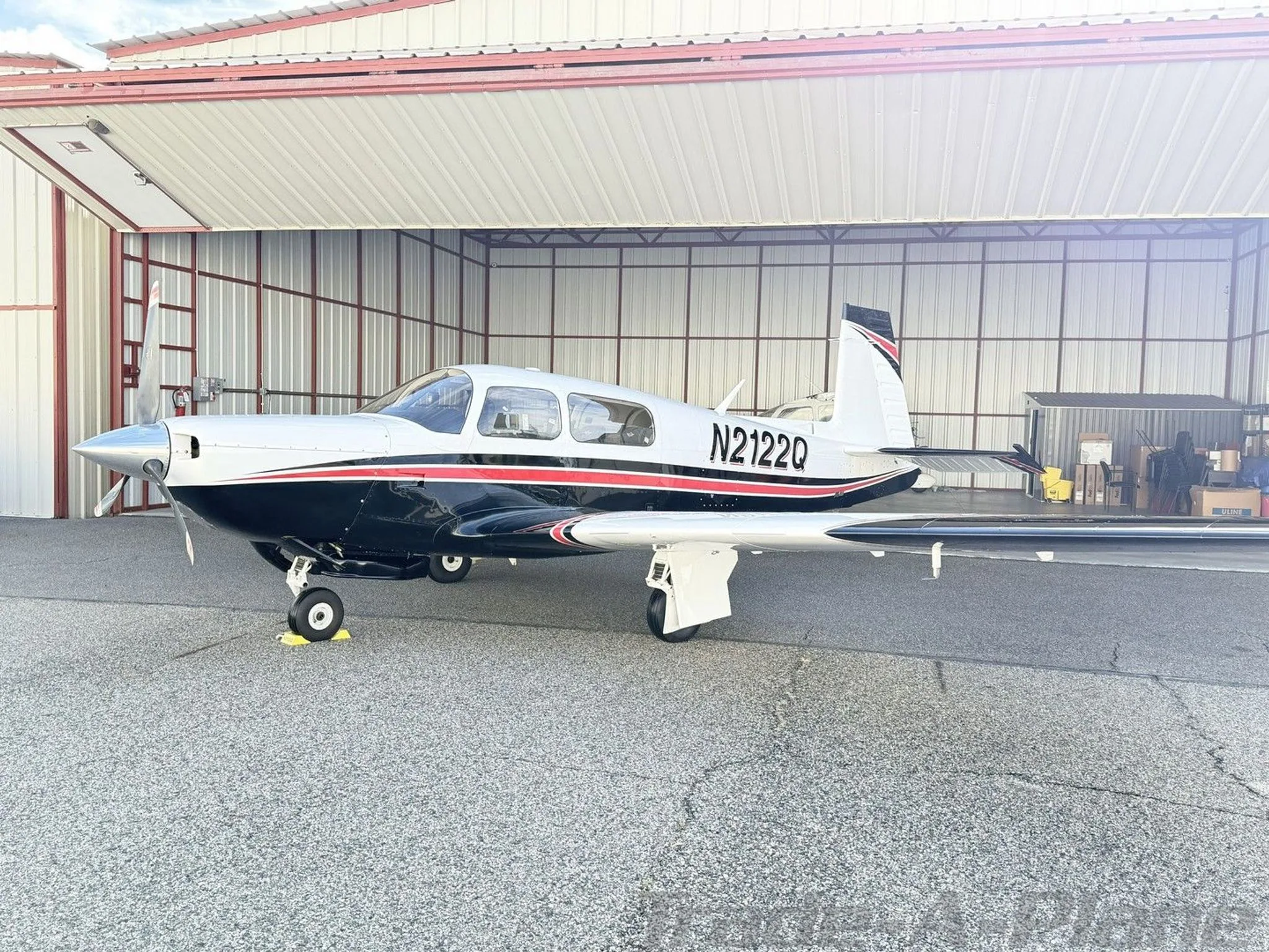 A white and black Mooney M20K aircraft with red stripes, registration N2122Q, parked in front of an open hangar.